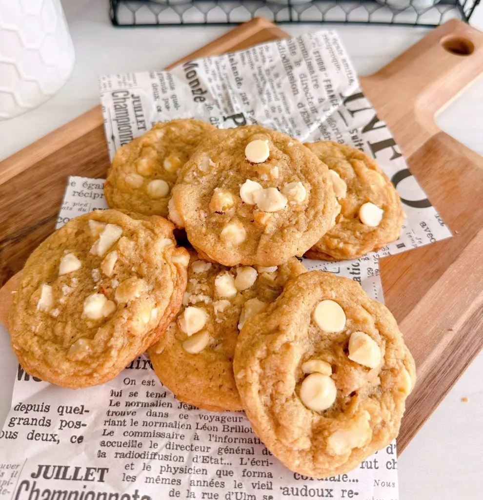 White Chocolate Chip Macadamia Nut cookies in a pile on a cutting board with news paper print.