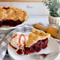 A slice of blackberry pie with a scoop of vanilla ice cream on a white plate with the whole pie in the background.