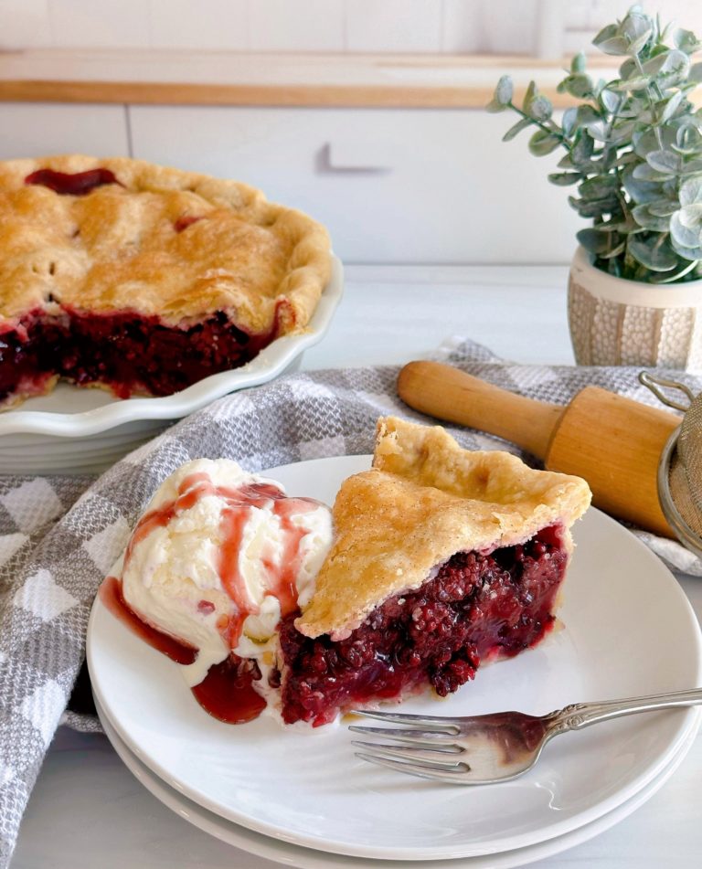 A slice of blackberry pie with a scoop of vanilla ice cream on a white plate with the whole pie in the background.