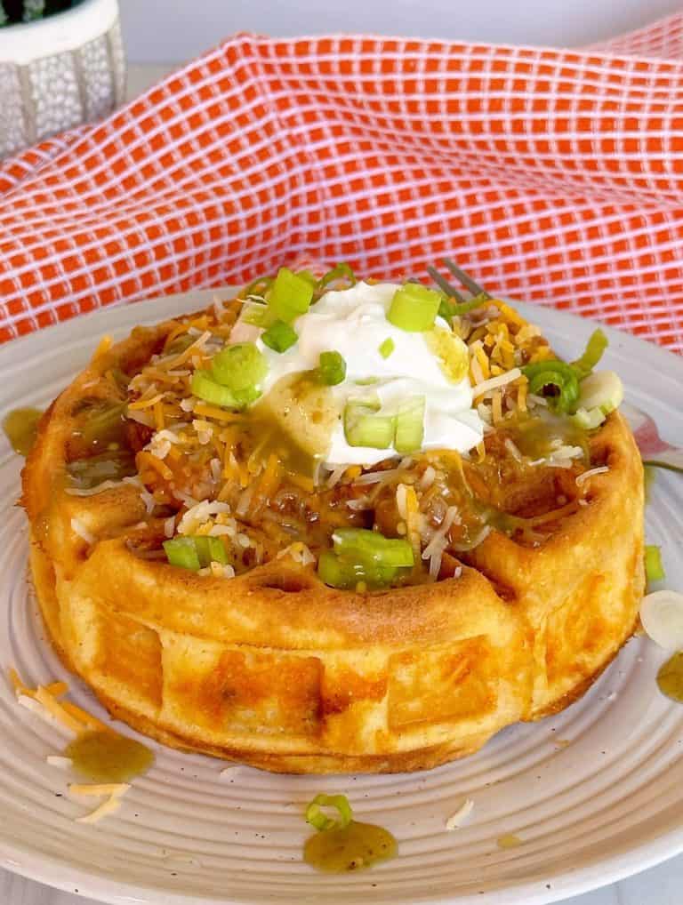 Cornbread waffle stuffed with chili and cheese and topped with sour cream and more cheese on a dinner plate.