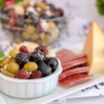 Marinated Olives with feta on a cheese board with a larger bowl in the background.
