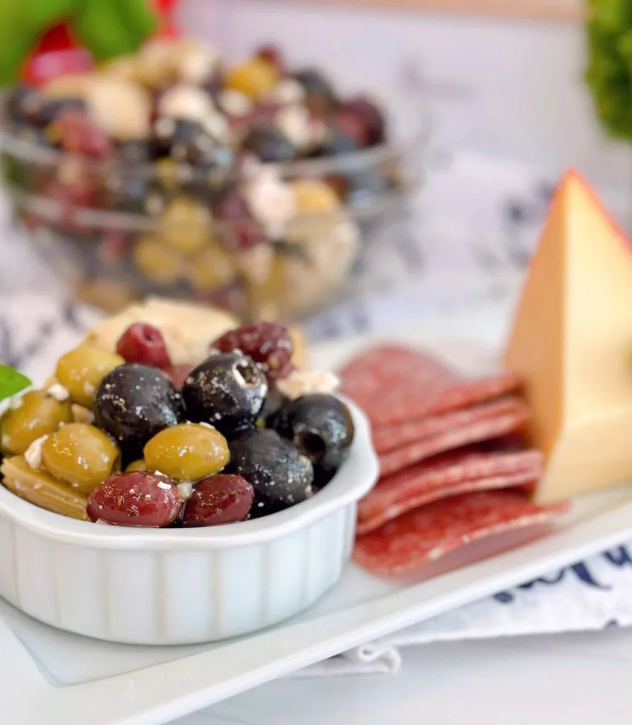 Marinated Olives with feta on a cheese board with a larger bowl in the background.
