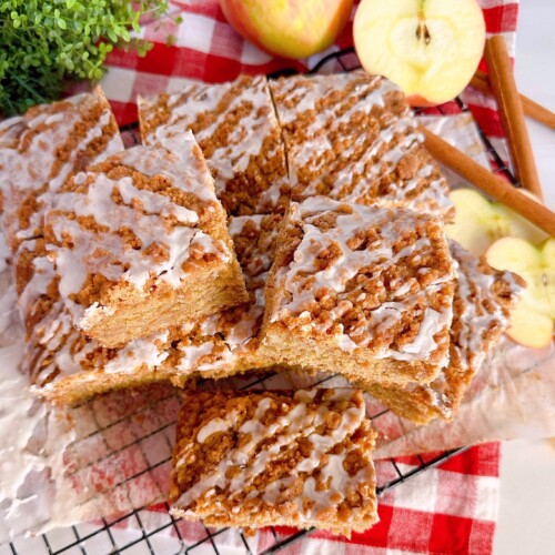 Apple Cider Crumb Cake cut and staked on a cooling rack surrounded by fresh apples and cinnamon sticks.