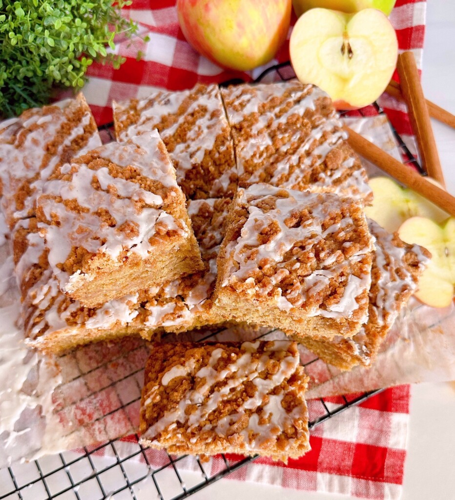 Apple Cider Crumb Cake cut and staked on a cooling rack surrounded by fresh apples and cinnamon sticks.