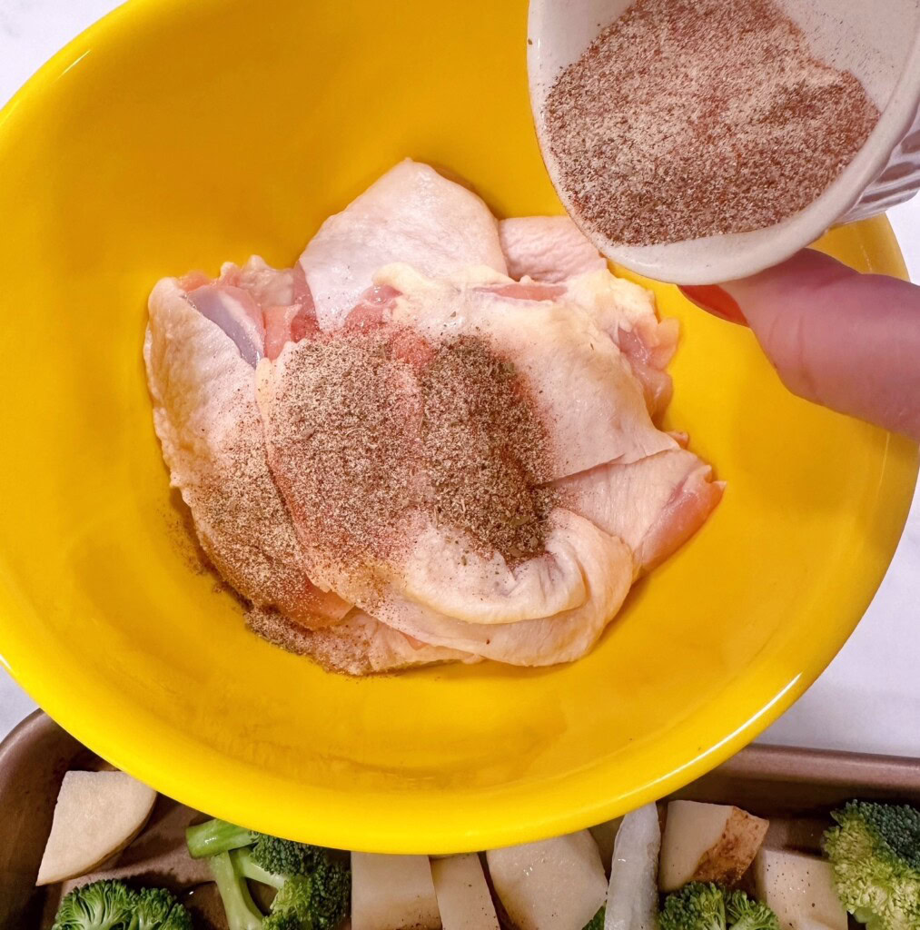 Chicken thighs in a bowl, with seasonings being sprinkled on top.