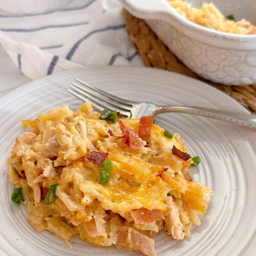 A serving of chicken hash brown casserole on a dish with a fork. The full casserole dish in the background.