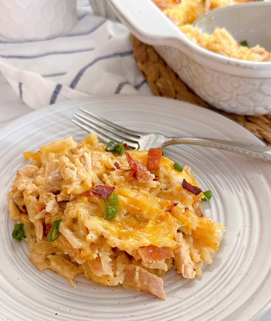 A serving of chicken hash brown casserole on a dish with a fork. The full casserole dish in the background.