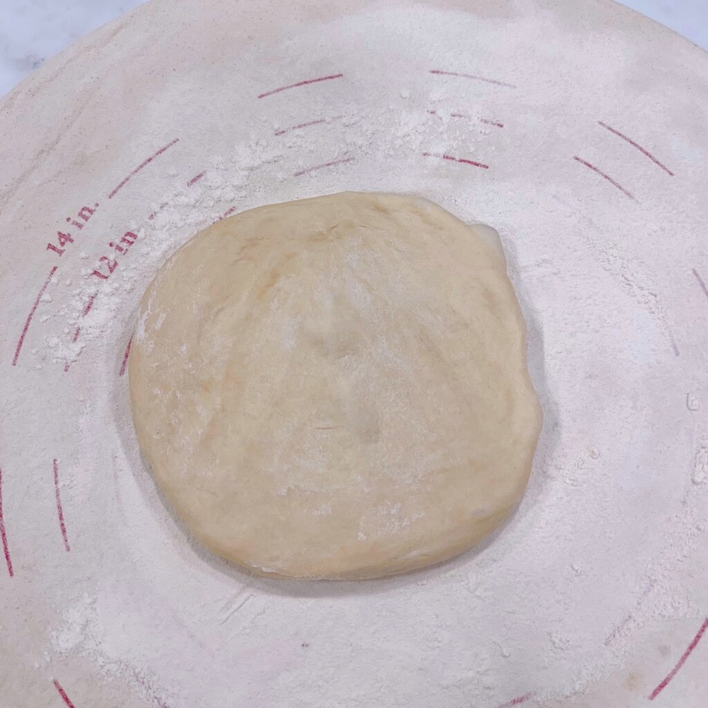Pizza dough pressed into a flat disc on a lightly floured surface.