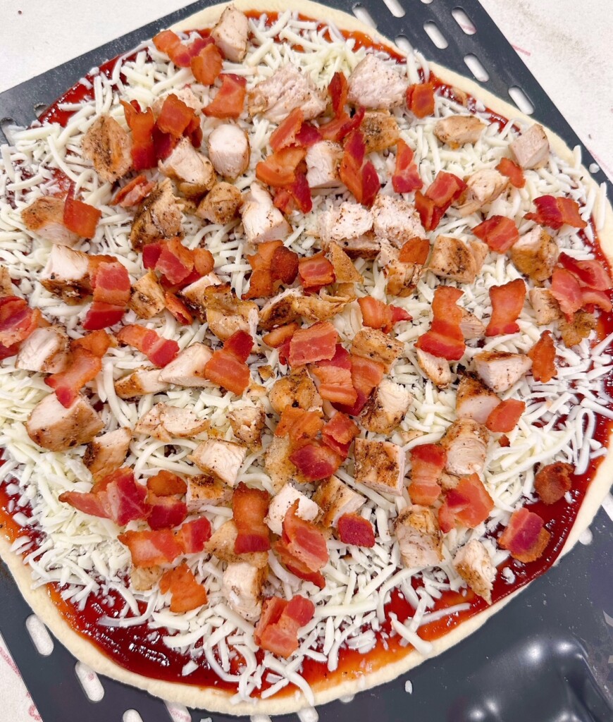 Chicken and bacon sprinkled over the top of the cheese on the pizza.